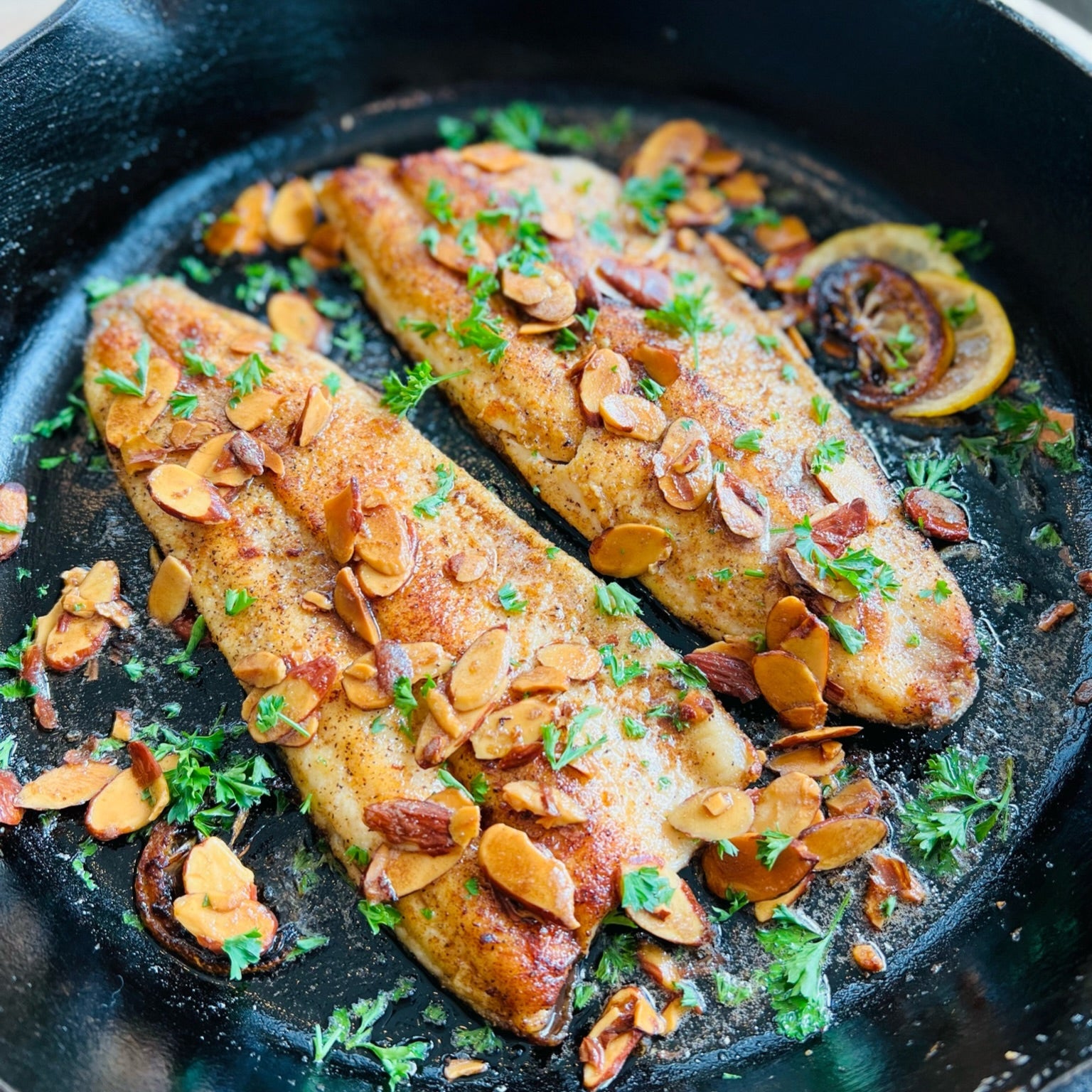 Two pieces of fish with almond flakes and herbs in a black cast iron skillet.