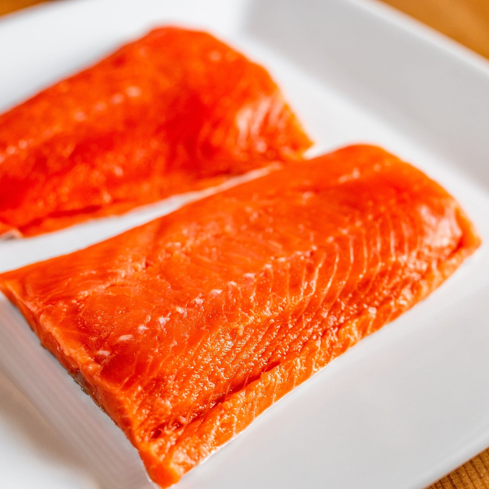 Two fresh salmon fillets on a white plate, displaying a bright orange color and skin-on texture.